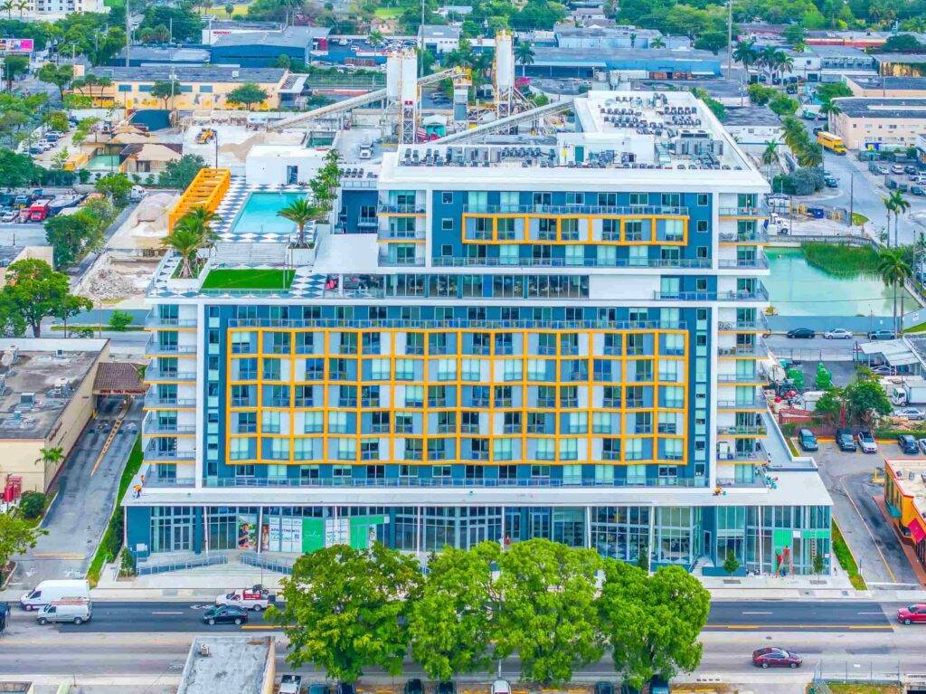Aerial view of a modern, multi-story building with colorful geometric patterns, surrounded by city streets and green spaces.