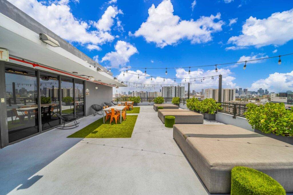 Modern rooftop patio with seating, city skyline views, and vibrant greenery under a bright blue sky.