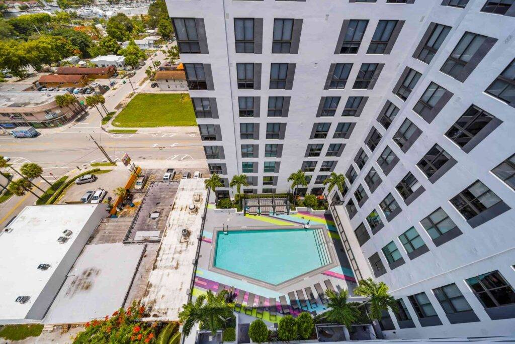 Aerial view of a modern urban apartment complex with a colorful rooftop pool and surrounding palm trees.