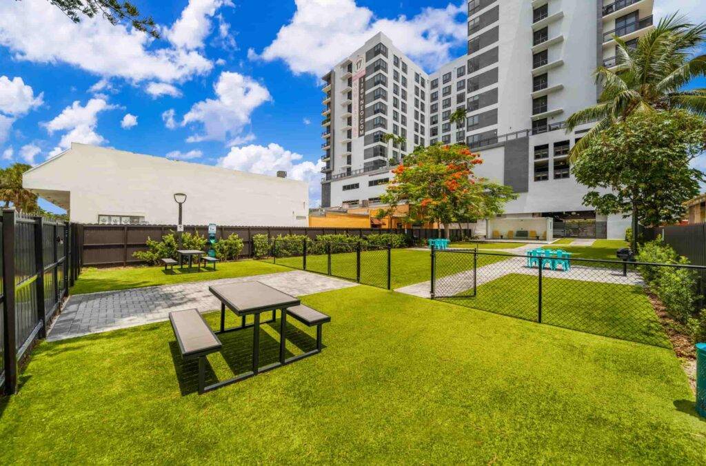 Urban park with picnic benches, greenery, and tall buildings under a blue sky with clouds.