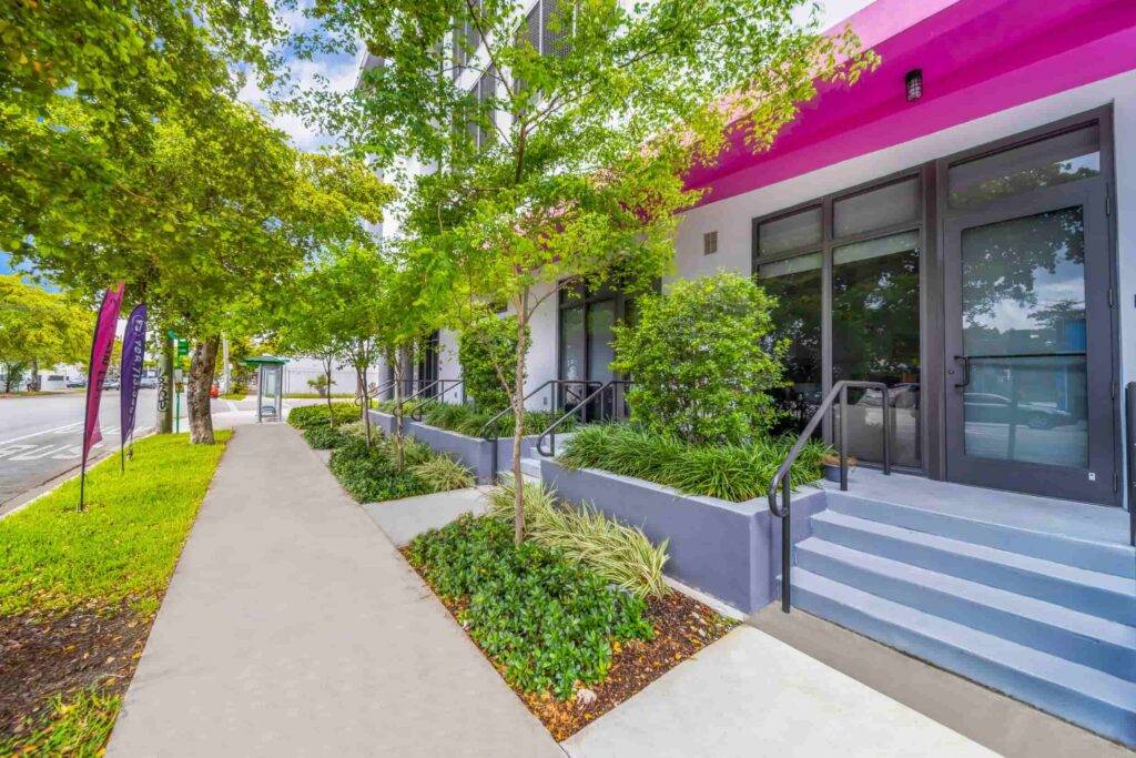 Vibrant street view of a modern building with pink accents surrounded by lush greenery.