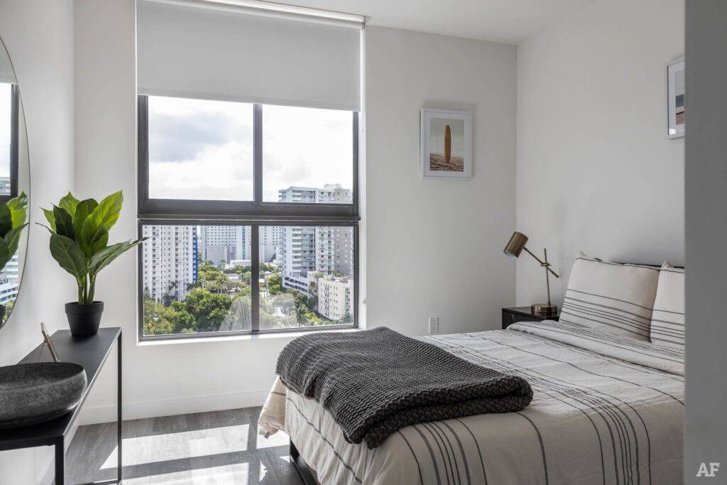 Modern bedroom with large window view of cityscape, featuring cozy bed and potted plant.