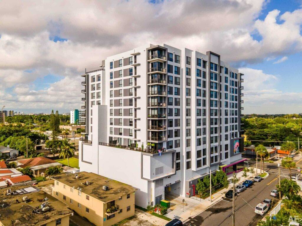 Modern apartment building in sunny cityscape with lush greenery and dramatic skies.