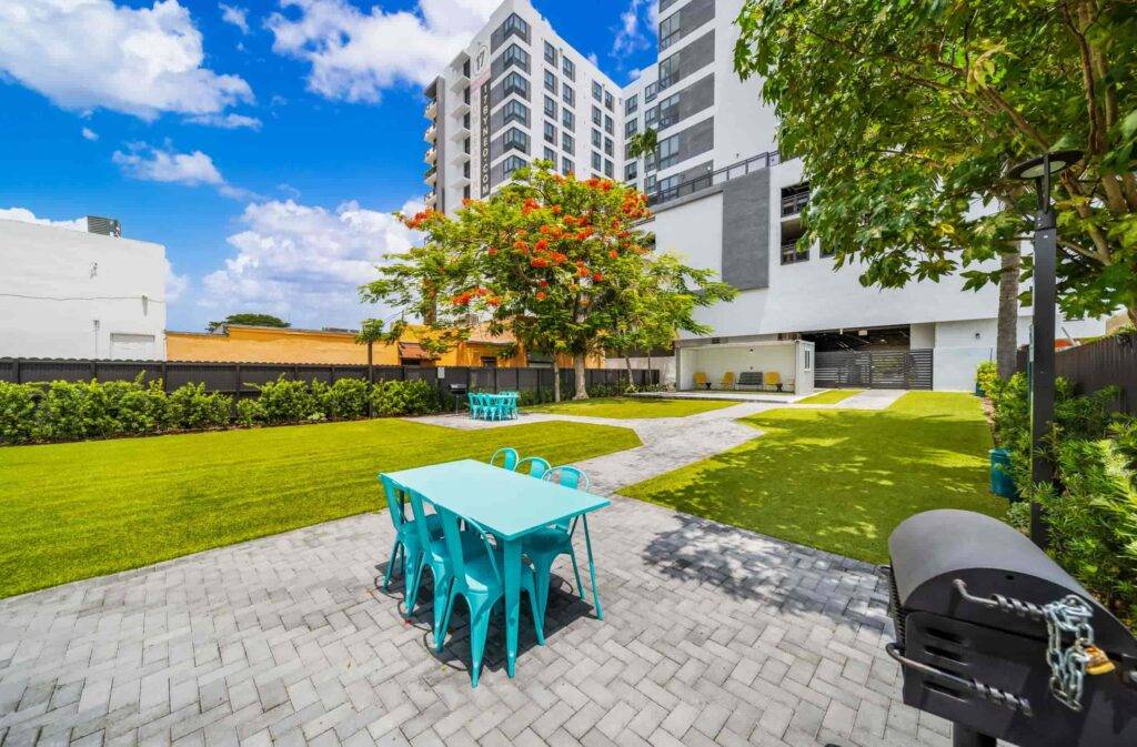 Modern urban courtyard with turquoise tables, lush greenery, and a towering apartment building under a blue sky.