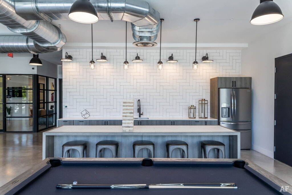 Modern industrial kitchen with sleek bar stools, pool table in foreground, and geometric backsplash.