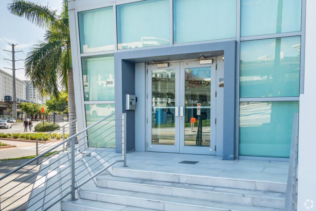 Modern building entrance with glass facade and stainless steel railings in Coral Gables, FL, featuring palm trees and urban surroundings.