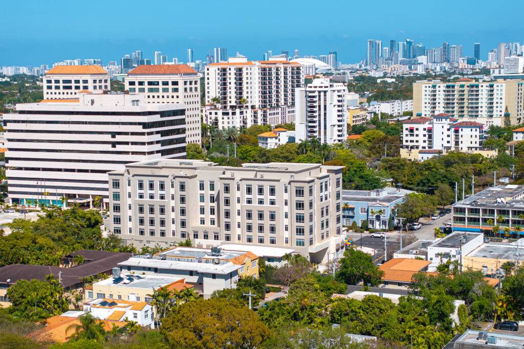 Aerial view of Coral Gables with modern buildings, lush greenery, and a distant city skyline under a clear blue sky.