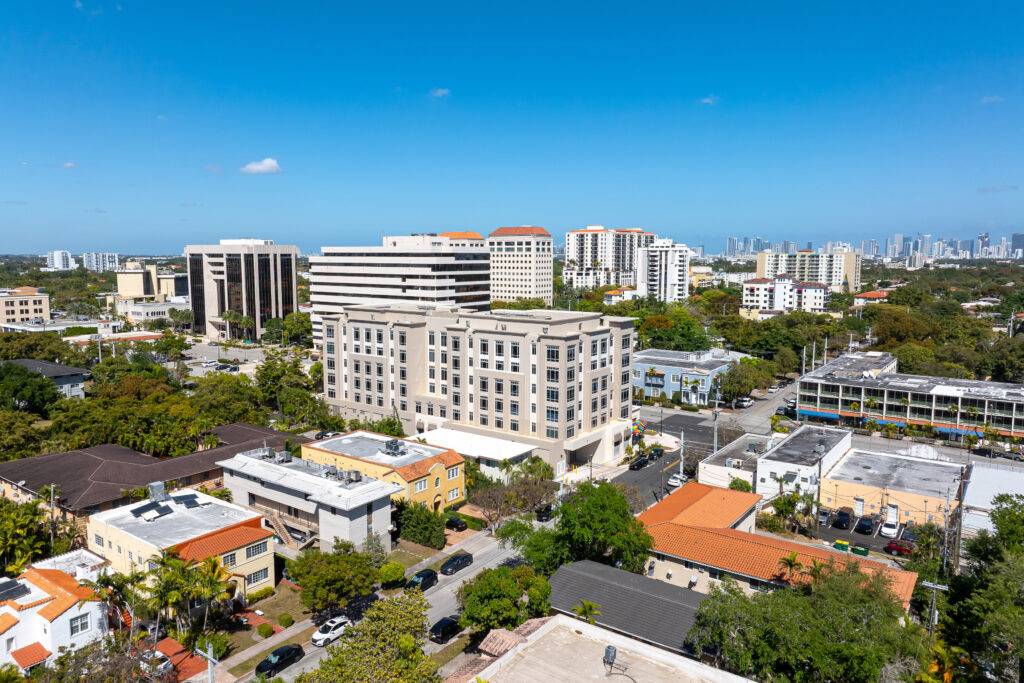Aerial view of Coral Gables with modern buildings and lush greenery under a clear blue sky.