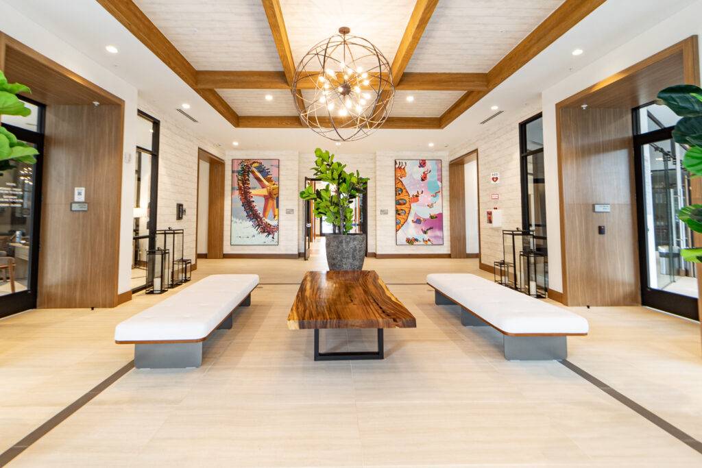 Modern lobby with wooden accents, white benches, abstract art, and a central plant arrangement under a stylish chandelier.