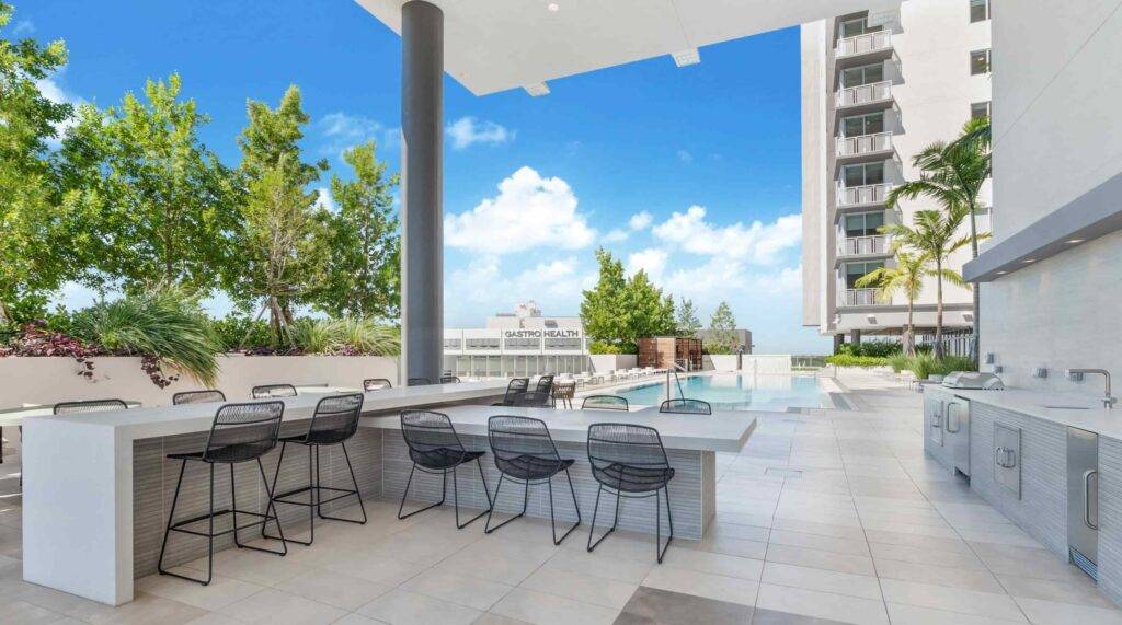 Modern outdoor kitchen area with bar stools and poolside view at a luxury building.