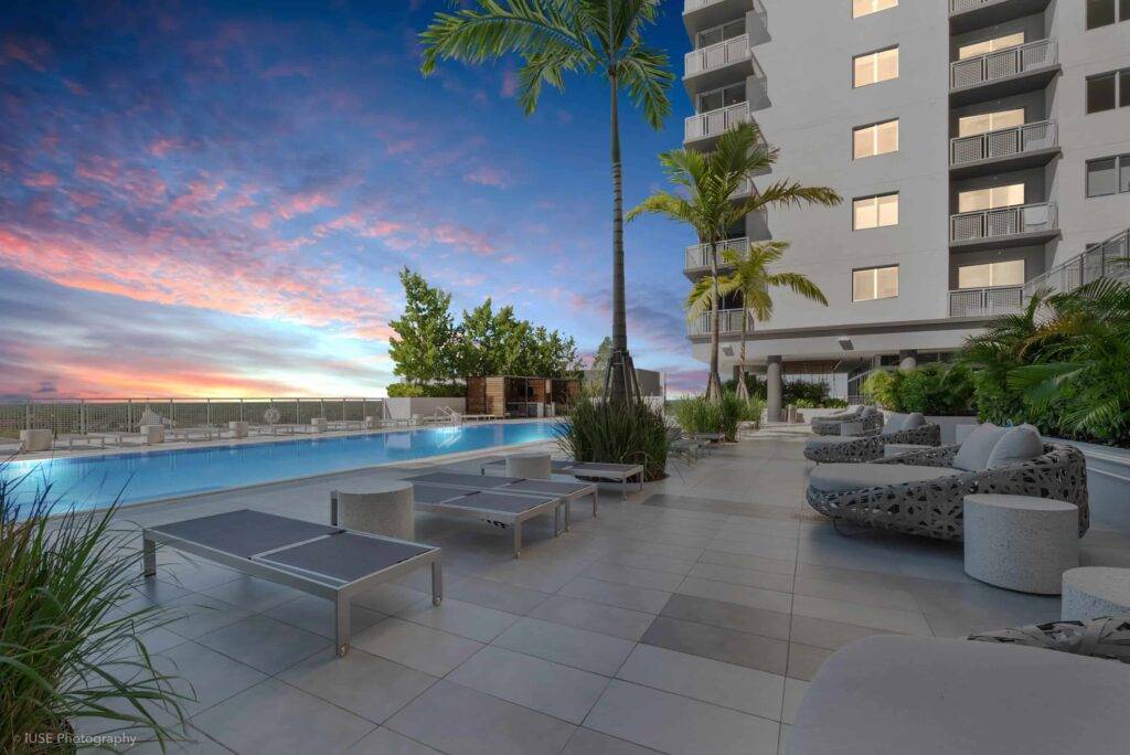 Modern rooftop pool terrace with sunset view, featuring sleek loungers and lush palm trees.