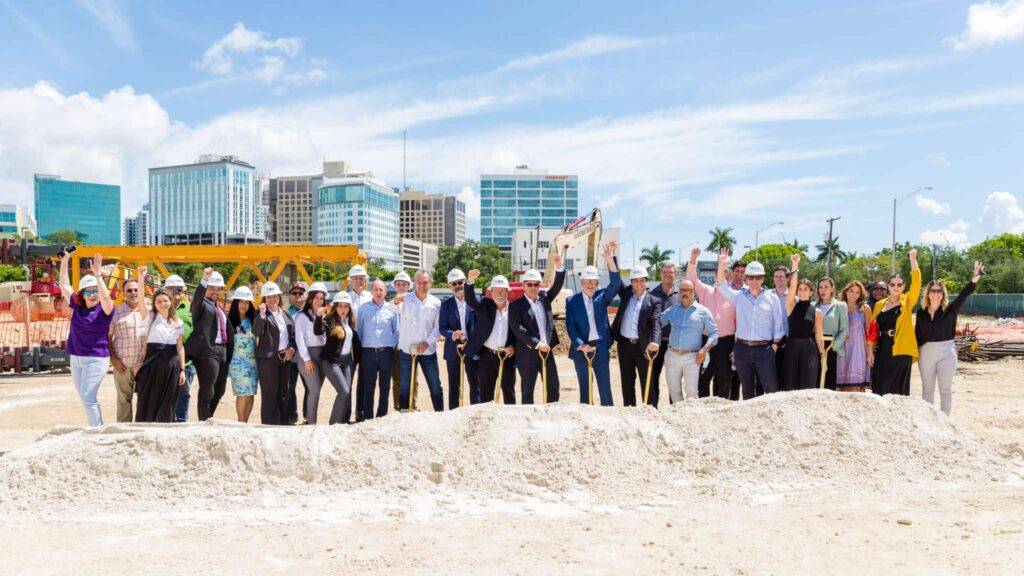 Group of people in hard hats celebrating a construction project at a sunny site with city buildings in the background.