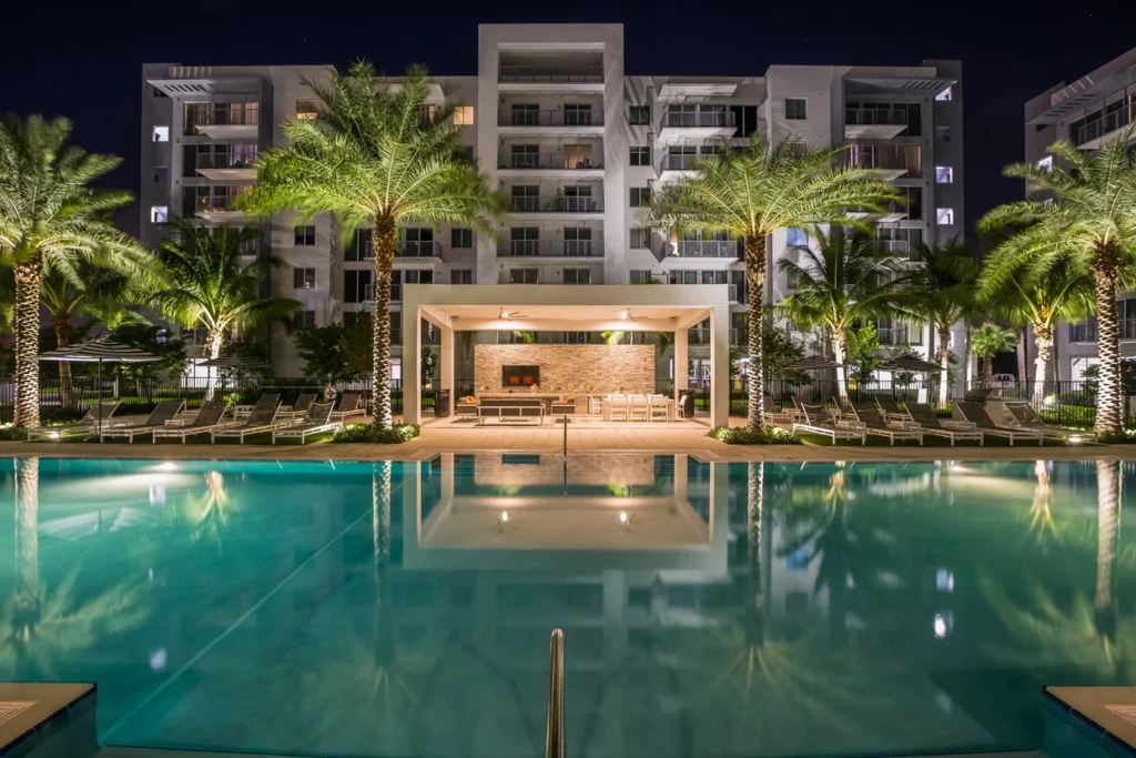 Nighttime view of luxury apartment complex with a large illuminated pool and palm trees.