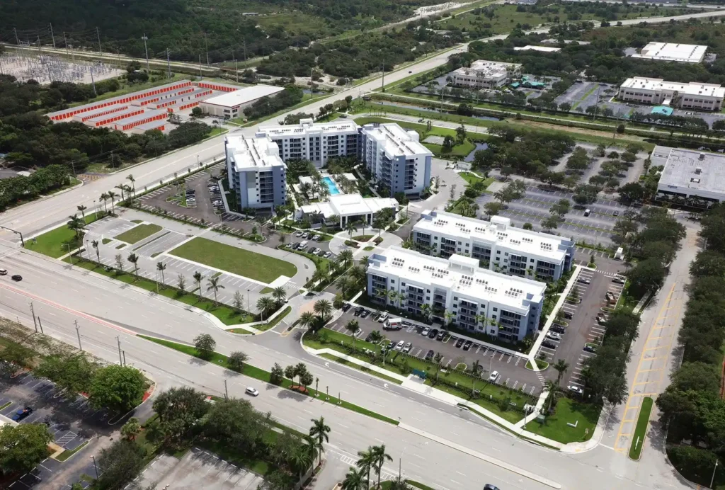 Aerial view of a modern residential complex surrounded by lush greenery and expansive parking areas.