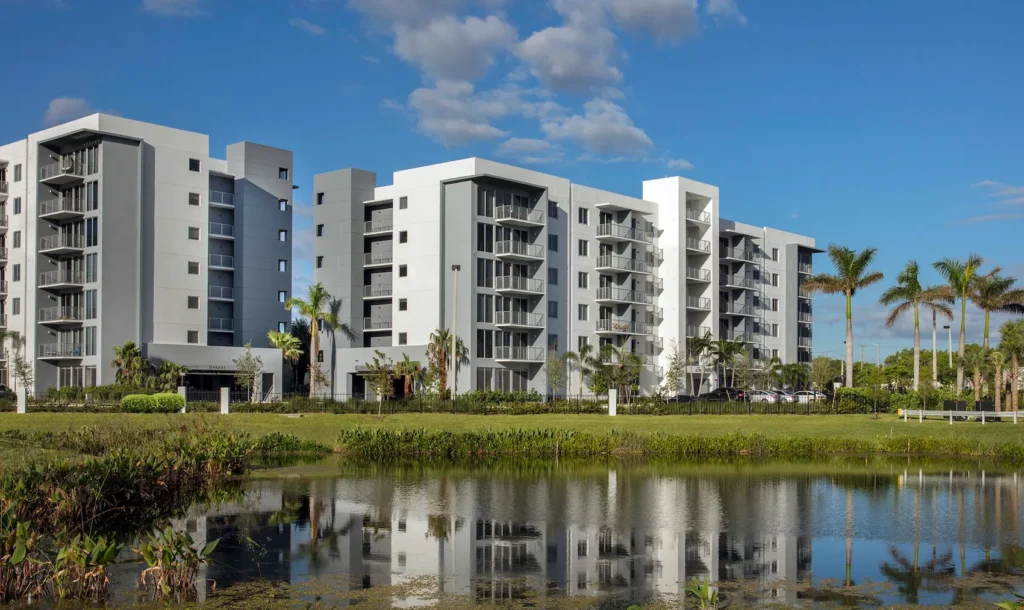 Modern apartment buildings beside a tranquil pond under a clear blue sky.