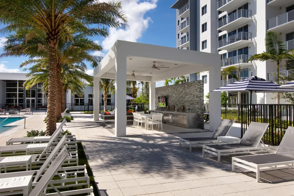 Modern outdoor lounge area with lounge chairs, a shaded pergola, and tall palm tree by an apartment building.