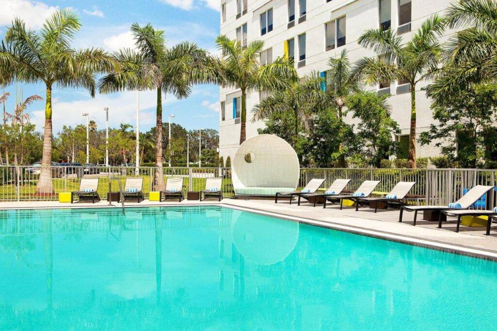 Modern hotel poolside with lounge chairs and palm trees under a clear blue sky.