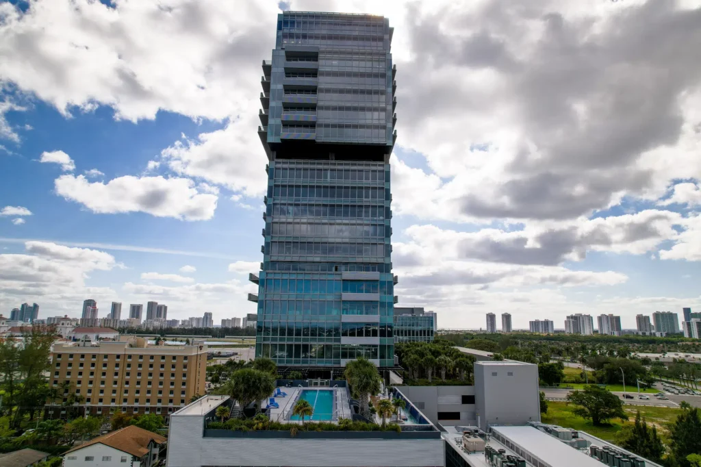 Modern skyscraper with a unique design against a cloudy sky, surrounded by urban landscape and lush greenery.