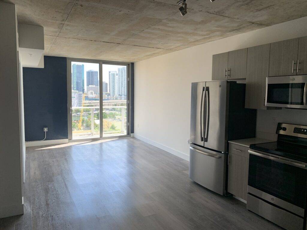 Modern apartment kitchen with stainless steel appliances and large window view of city skyline.