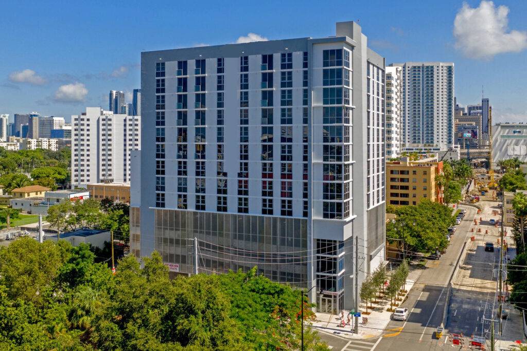 Modern high-rise building in a bustling cityscape with lush greenery and clear blue sky.