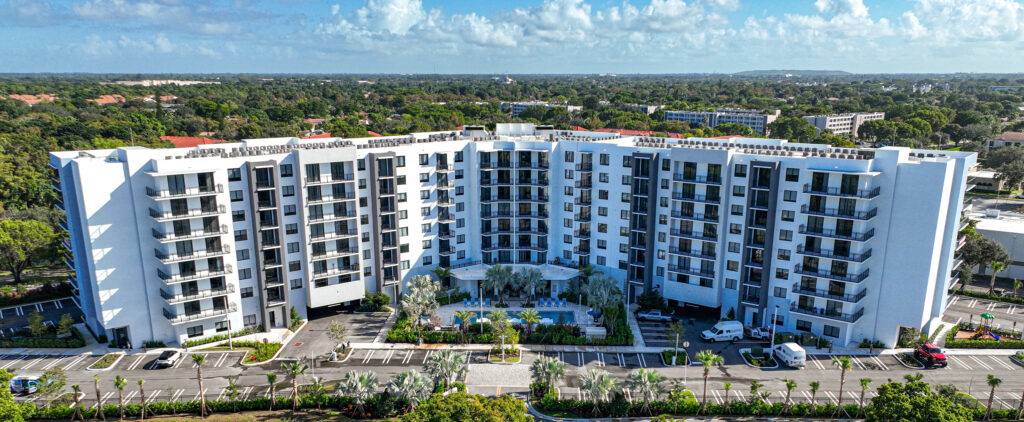 Aerial view of a modern white apartment complex surrounded by lush greenery and a clear blue sky.