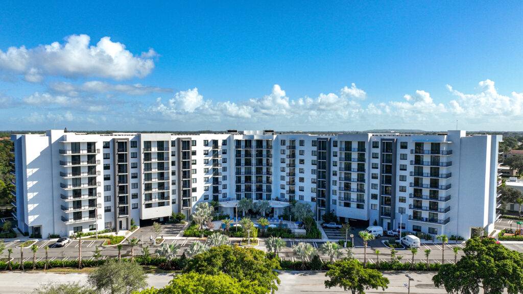 Aerial view of a modern white apartment building surrounded by lush greenery under a clear blue sky.