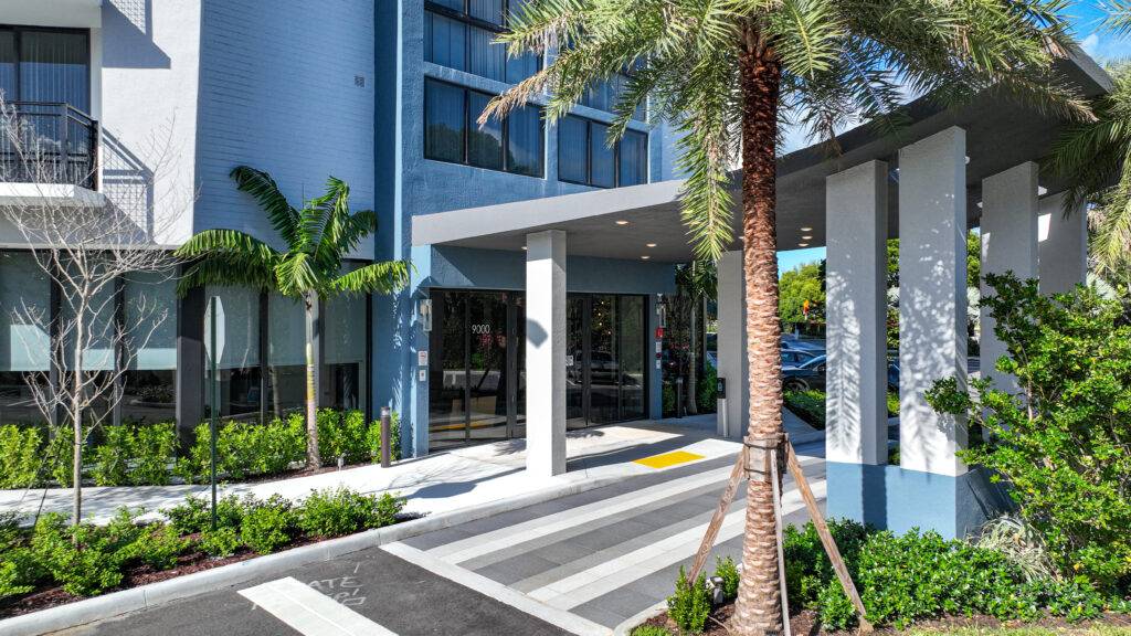 Modern hotel entrance with sleek architecture and tropical landscaping, featuring palm trees and lush greenery.
