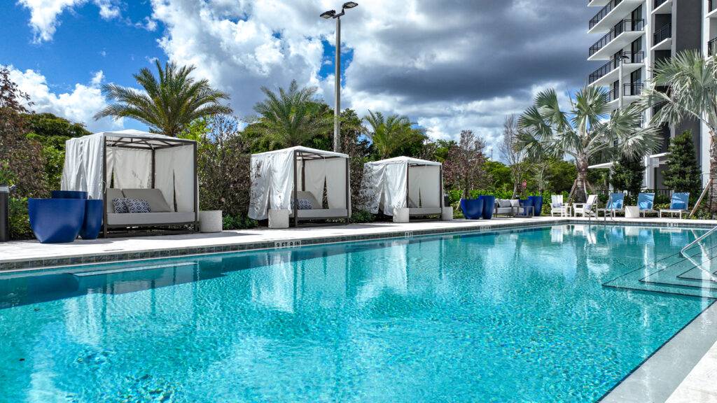 Luxurious outdoor pool with cabanas under palm trees and cloudy skies.