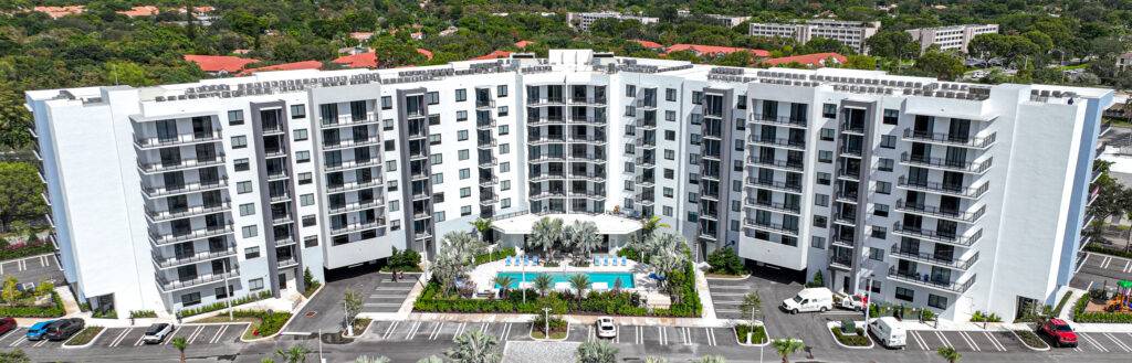 Aerial view of a modern white apartment complex with lush greenery and a central swimming pool.