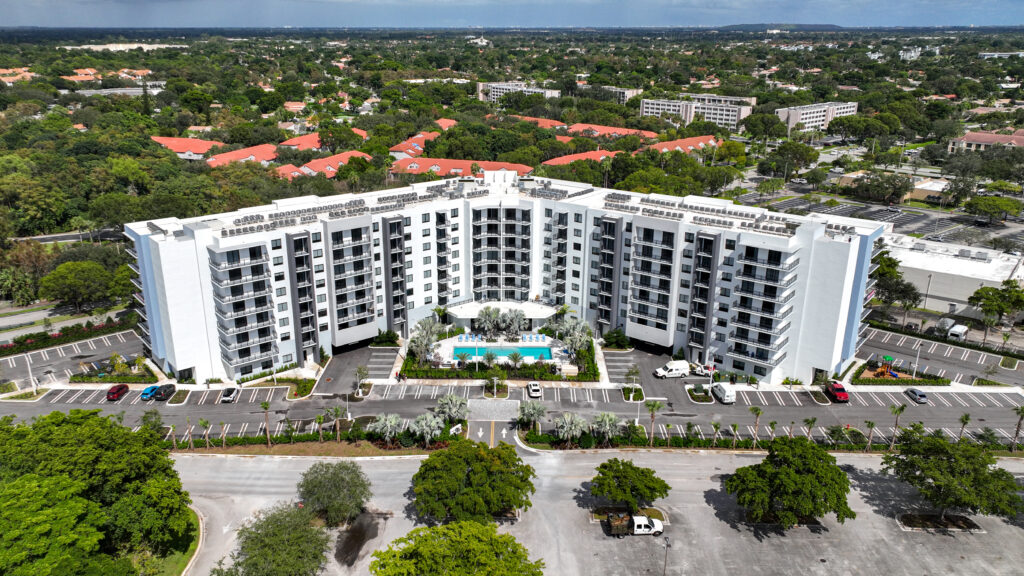 Aerial view of a modern white high-rise apartment complex with a central pool, surrounded by greenery and extensive parking.