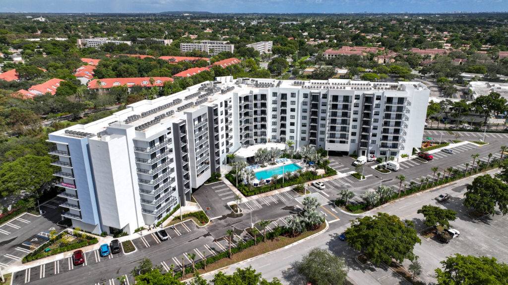 Aerial view of a modern white apartment complex with a central swimming pool surrounded by trees and parked cars.