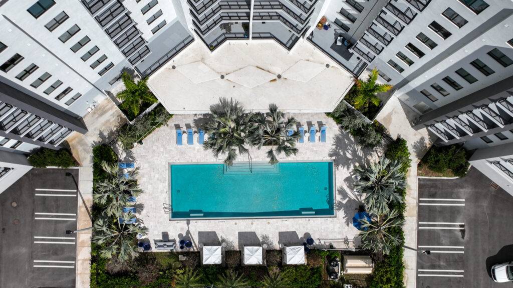 Aerial view of a modern apartment complex with a central turquoise pool surrounded by palm trees and sun loungers.