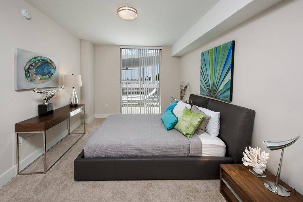 Modern bedroom with sleek gray bed, vibrant artwork, and natural light from window.