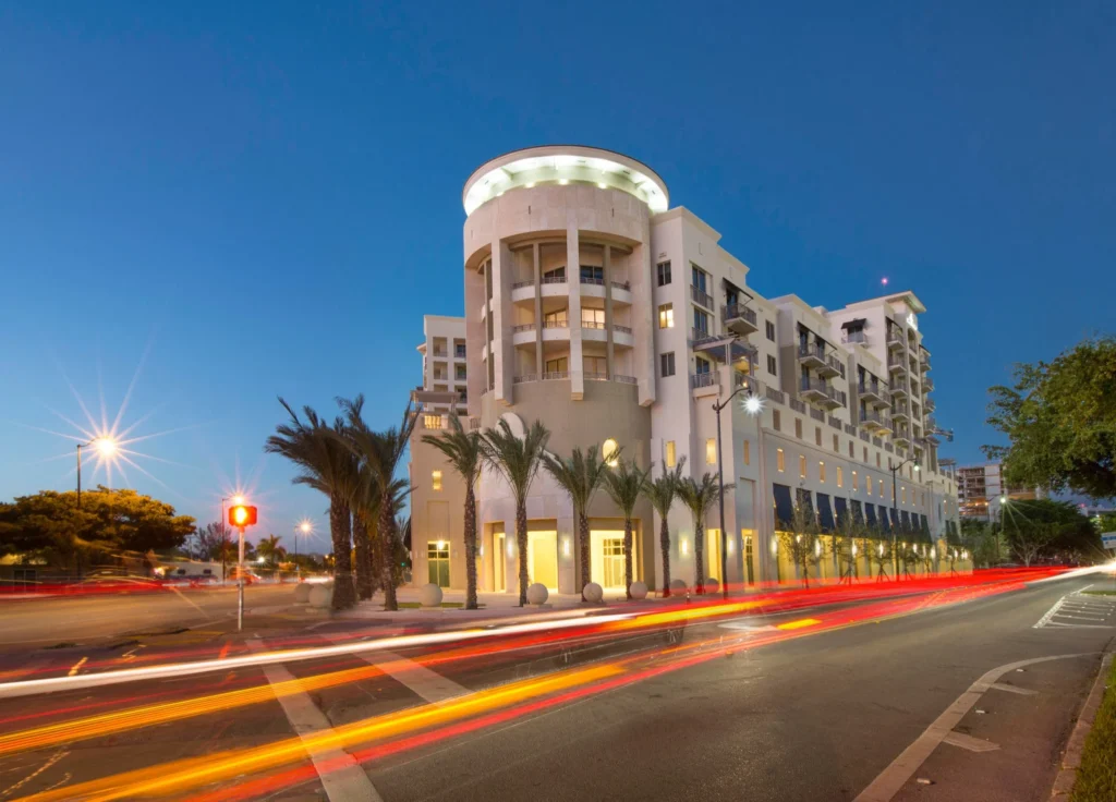 Modern multi-story building illuminated at dusk with palm trees and blurred traffic lights.