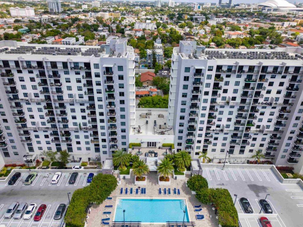 Aerial view of modern apartment complex with two tall buildings, a central courtyard, and a pool, surrounded by a bustling urban neighborhood.