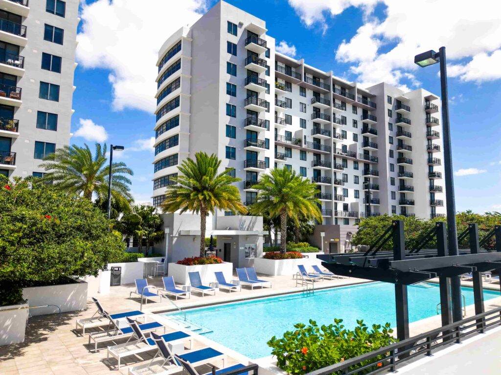 Modern apartment building with a sunny outdoor pool and lounge chairs, surrounded by lush greenery.