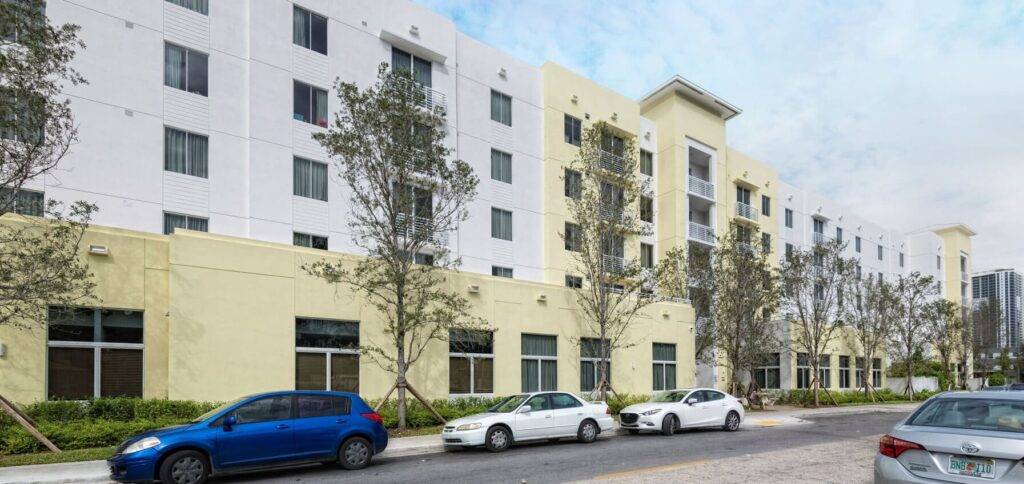 Modern apartment complex with yellow and white facade surrounded by trees and parked cars.