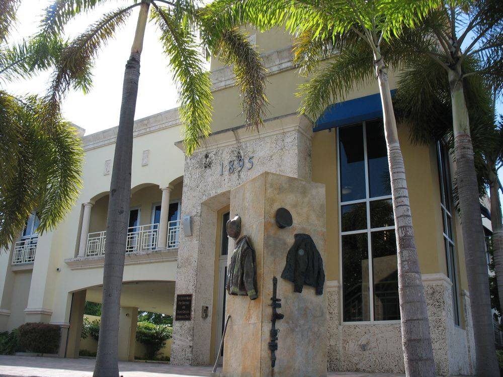 Modern building facade with palm trees and a stone sculpture in front.