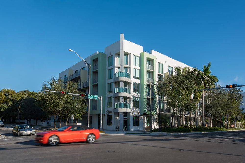 Modern apartment building at a busy urban intersection with a red convertible passing by.