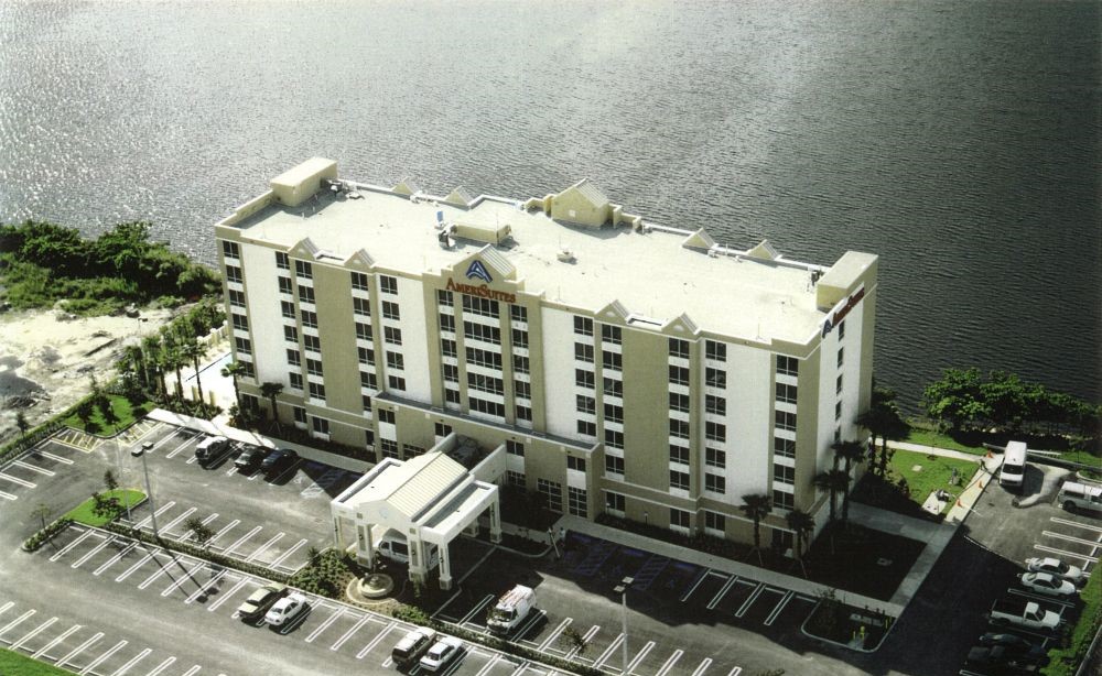 Aerial view of a multi-story hotel building beside a serene lake with parking area in front.