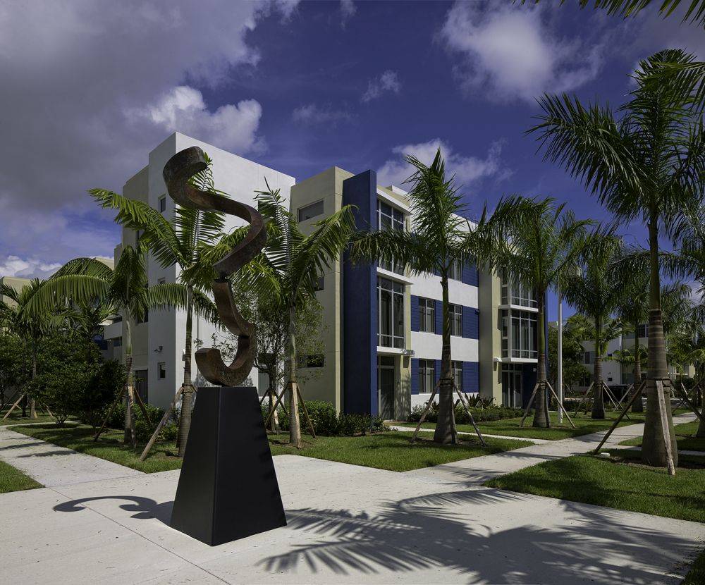 Modern art sculpture outside a contemporary building surrounded by palm trees under a partly cloudy sky.