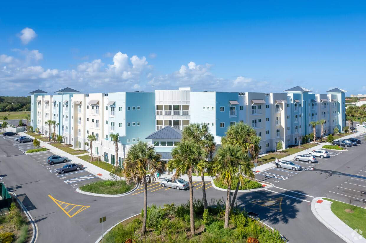 Modern apartment complex with pastel blue facade, palm trees, and a spacious parking area under a clear blue sky.