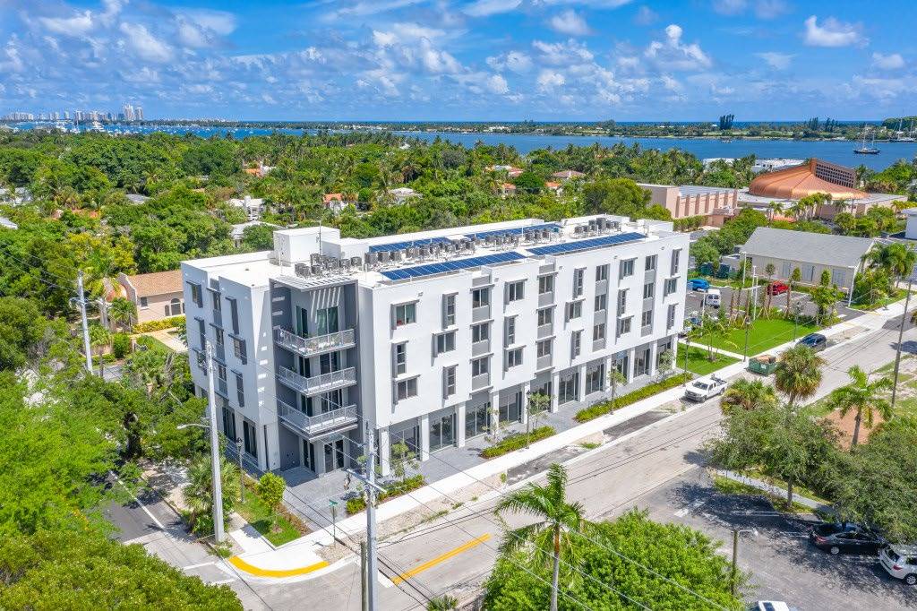 Aerial view of a modern white apartment building surrounded by lush greenery and a distant ocean backdrop.