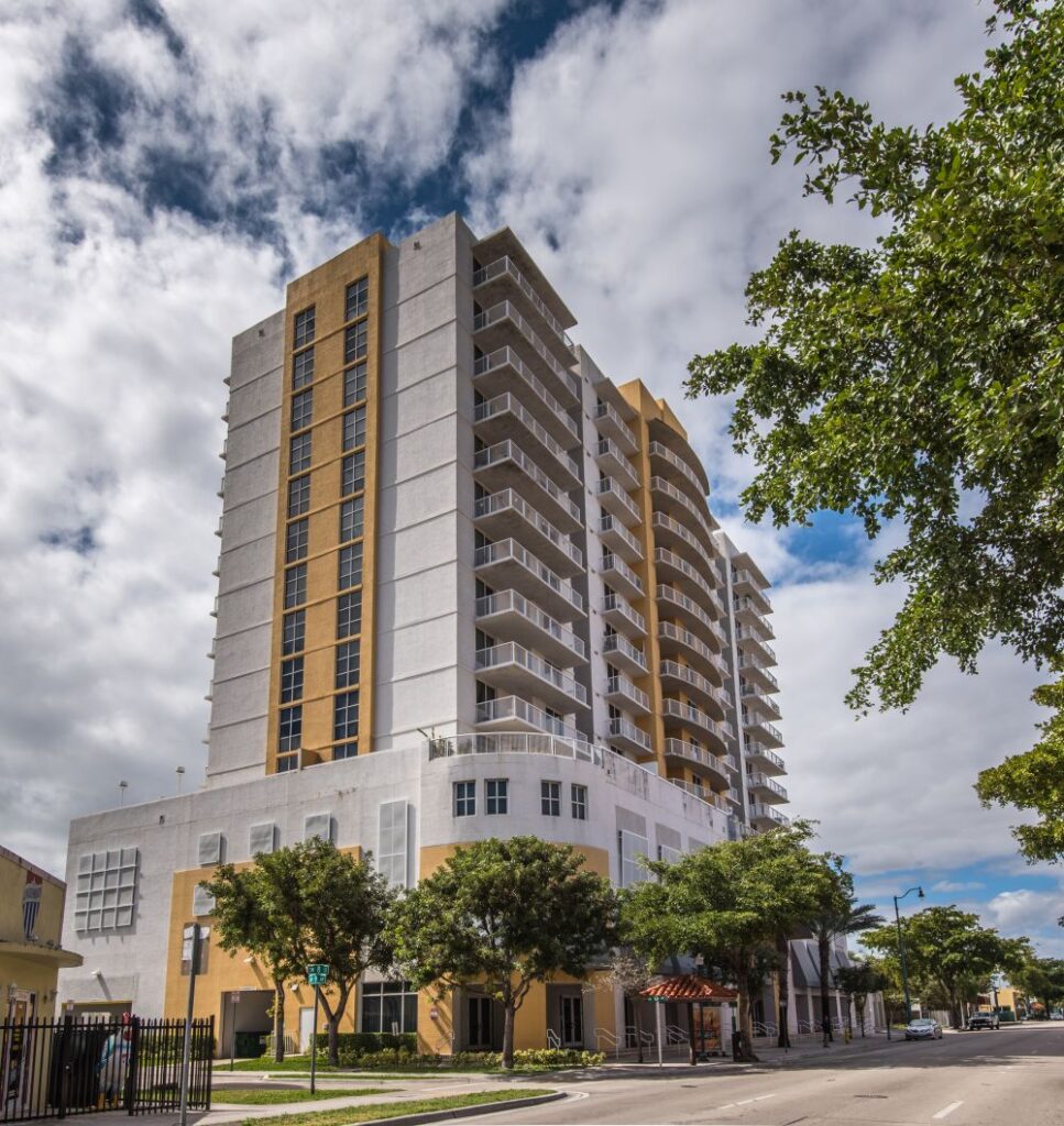 Modern high-rise building with a yellow and white facade under a cloudy sky, surrounded by lush green trees.