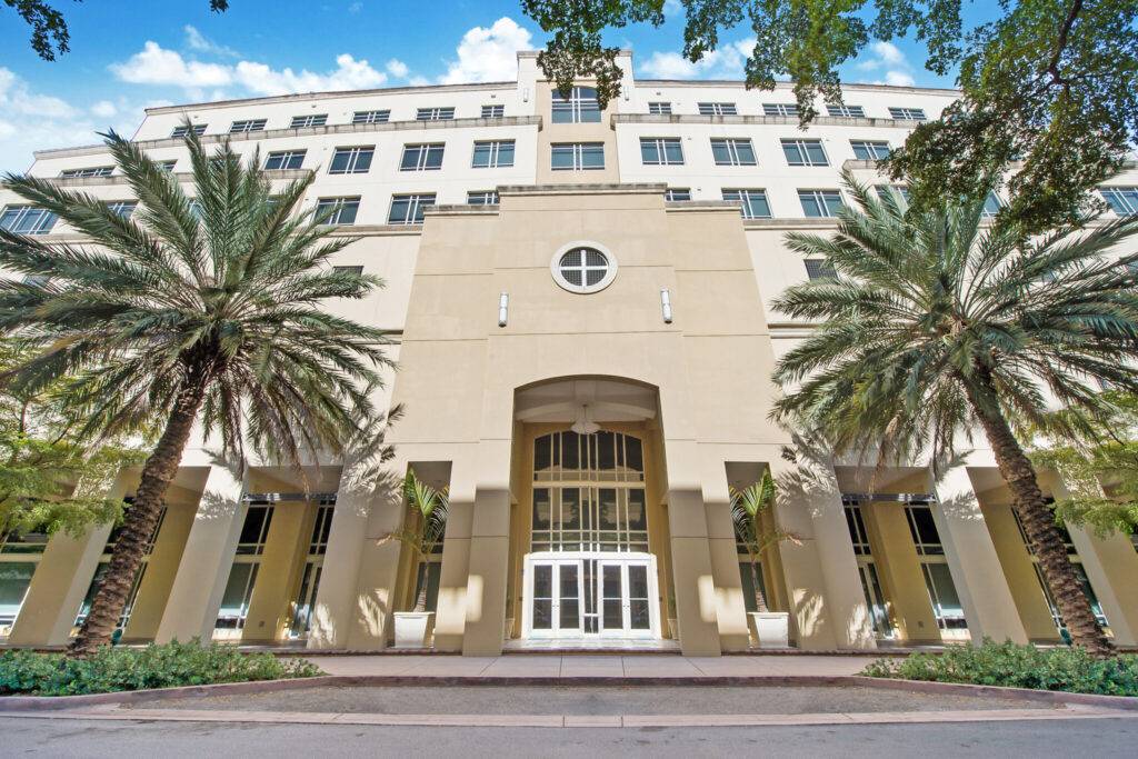 Elegant beige building with tall arched entrance and lush palm trees under a bright blue sky.
