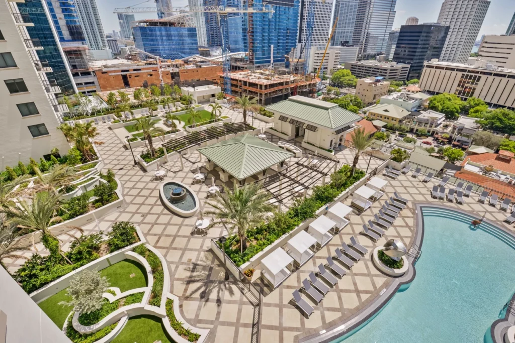 Aerial view of a modern urban resort with a pool, lush greenery, and skyscrapers in the background.