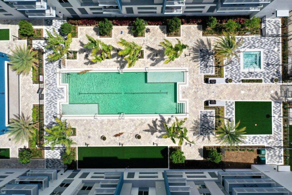 Aerial view of a modern apartment complex with a central swimming pool, lush palm trees, and surrounding green lawns.