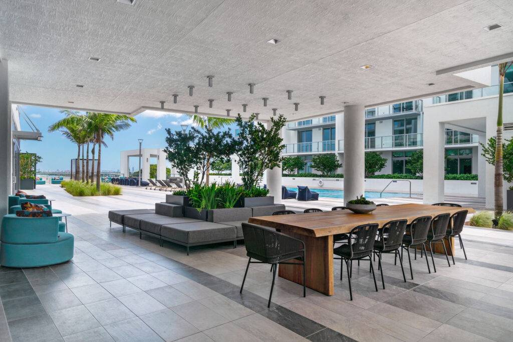 Outdoor lounge area with a wooden dining table, modern seating, greenery, and a view of a pool.