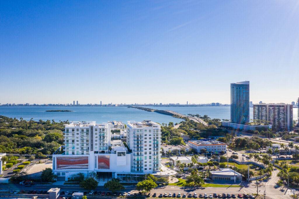Aerial view of modern waterfront cityscape with buildings, lush greenery, and a clear blue sky.
