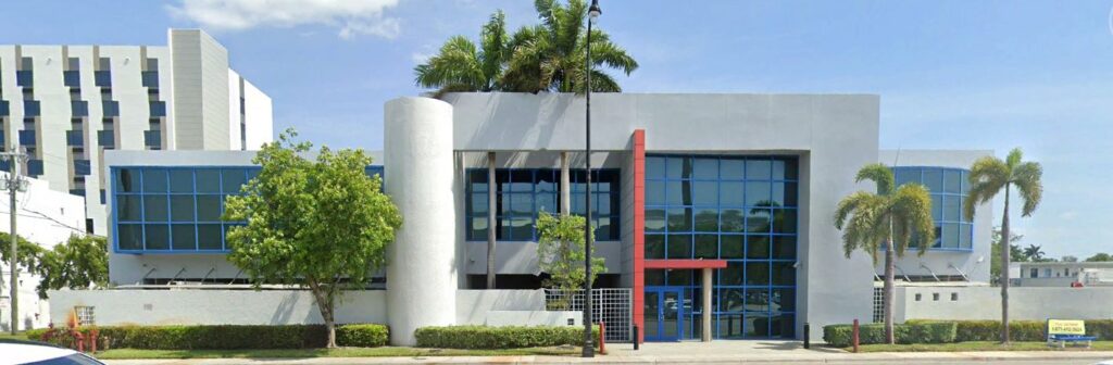 Modern building with tropical landscaping and large glass windows under a clear blue sky.